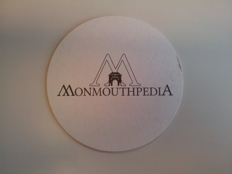 A round paper drink coaster printed with the Monmouthpedia logo; illustrative third-party example, not an Eco Friendly Boxes product.